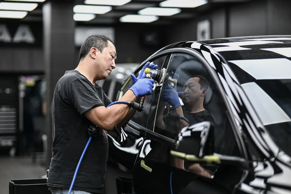 技師在黑色 Tesla 車側以研磨機進行玻璃拋光鍍膜,車窗映出施工區棋盤格天花板 誠美車體美容